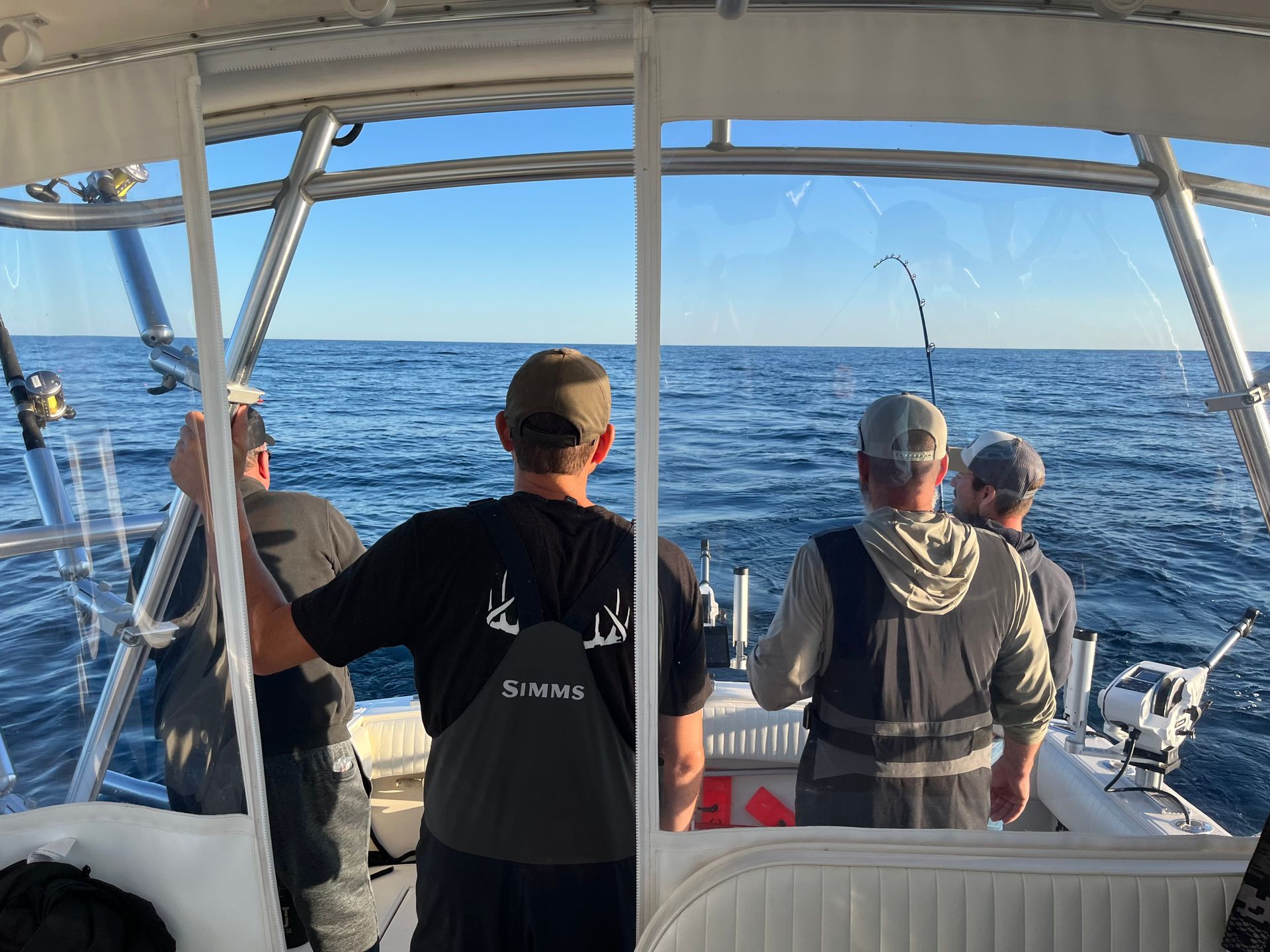 Men fishing from a boat on the ocean. One has a bent fishing rod. Clear skies and calm water.