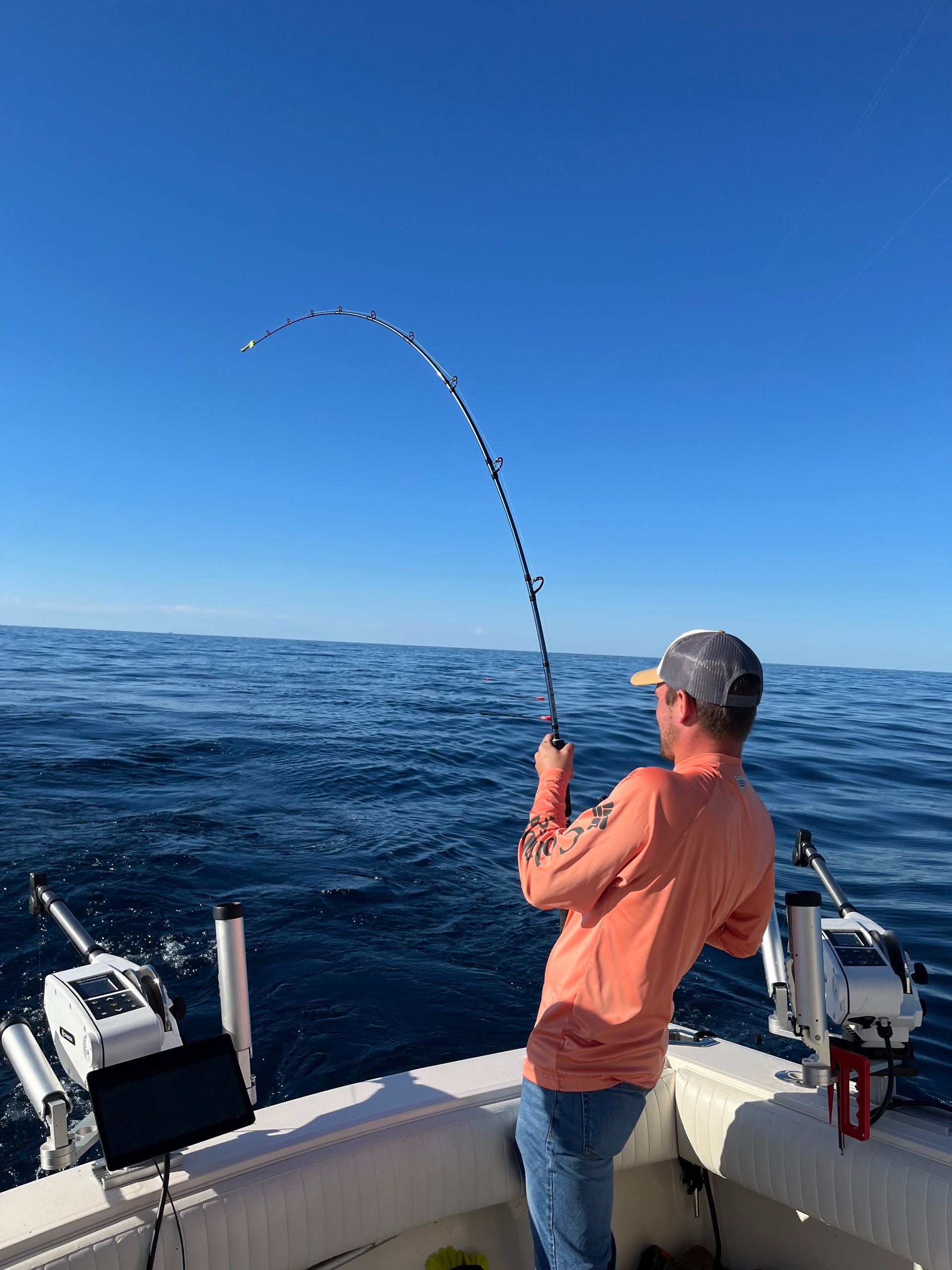 Man on boat reeling in a fish; bent fishing rod against blue sky and ocean.