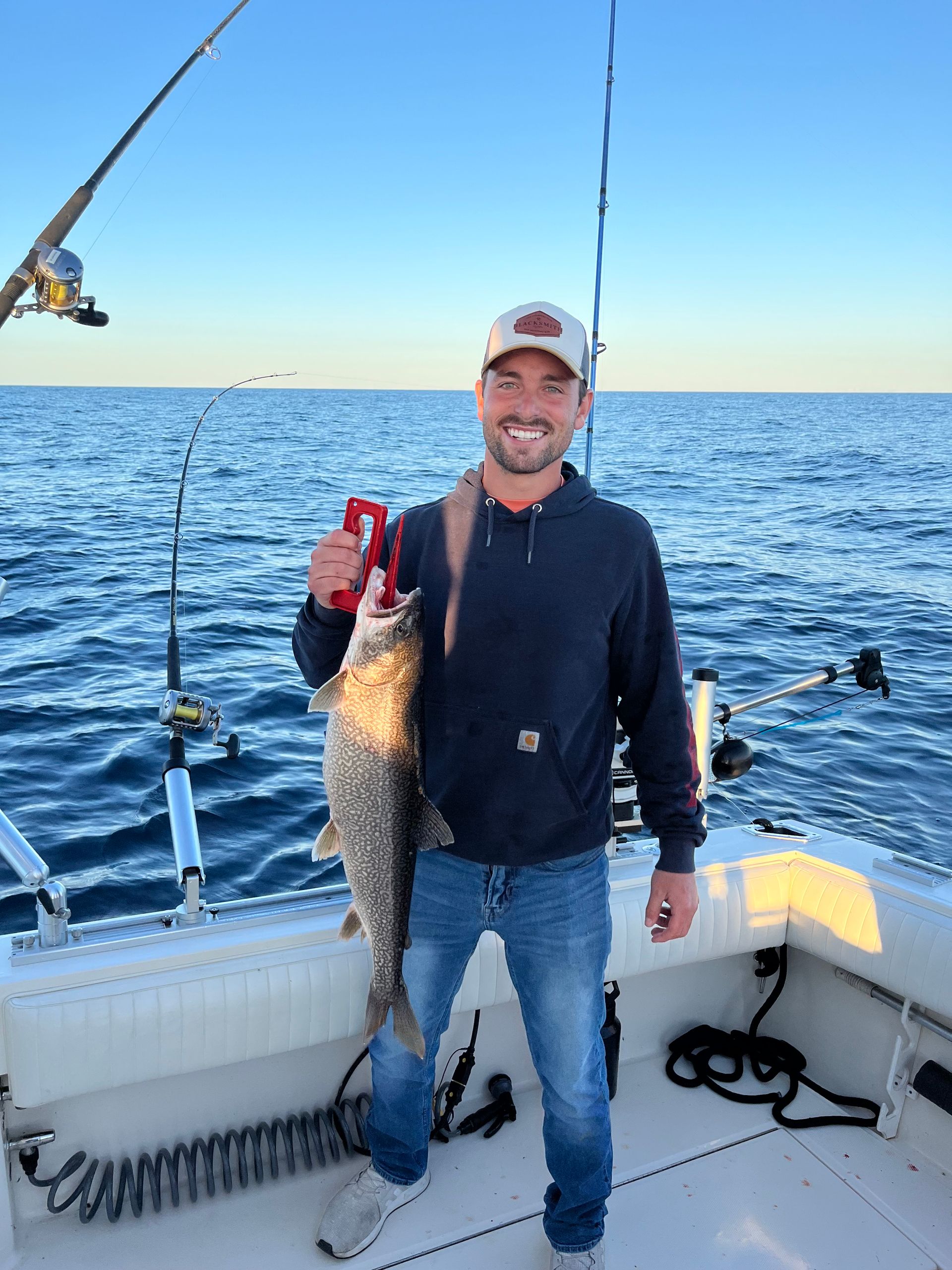 Man on a boat holding a fish, smiling. Blue jeans, navy hoodie, sunny day, water, fishing rods.