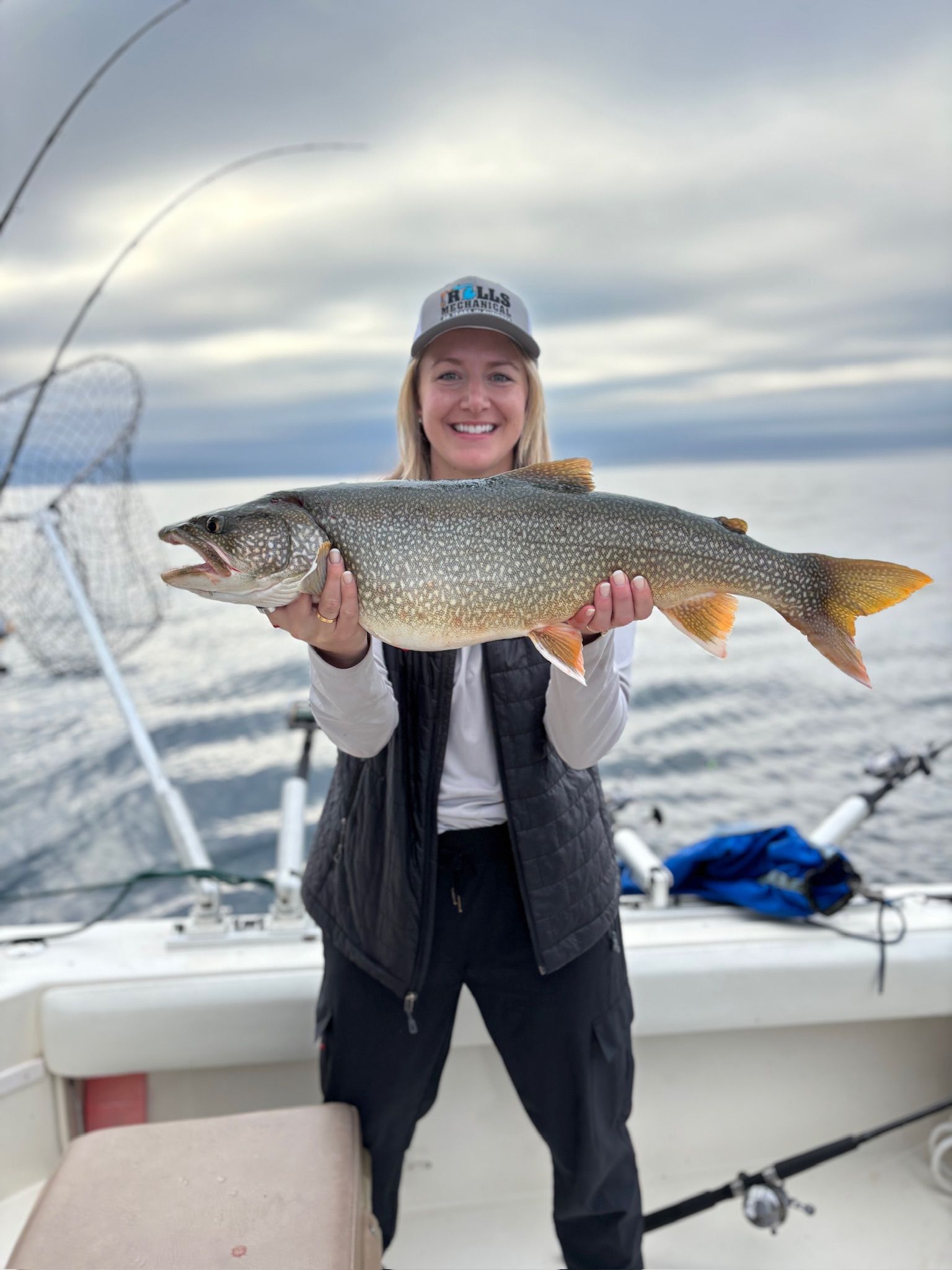 Woman smiling, holding a large fish on a boat. Cloudy sky, water visible.