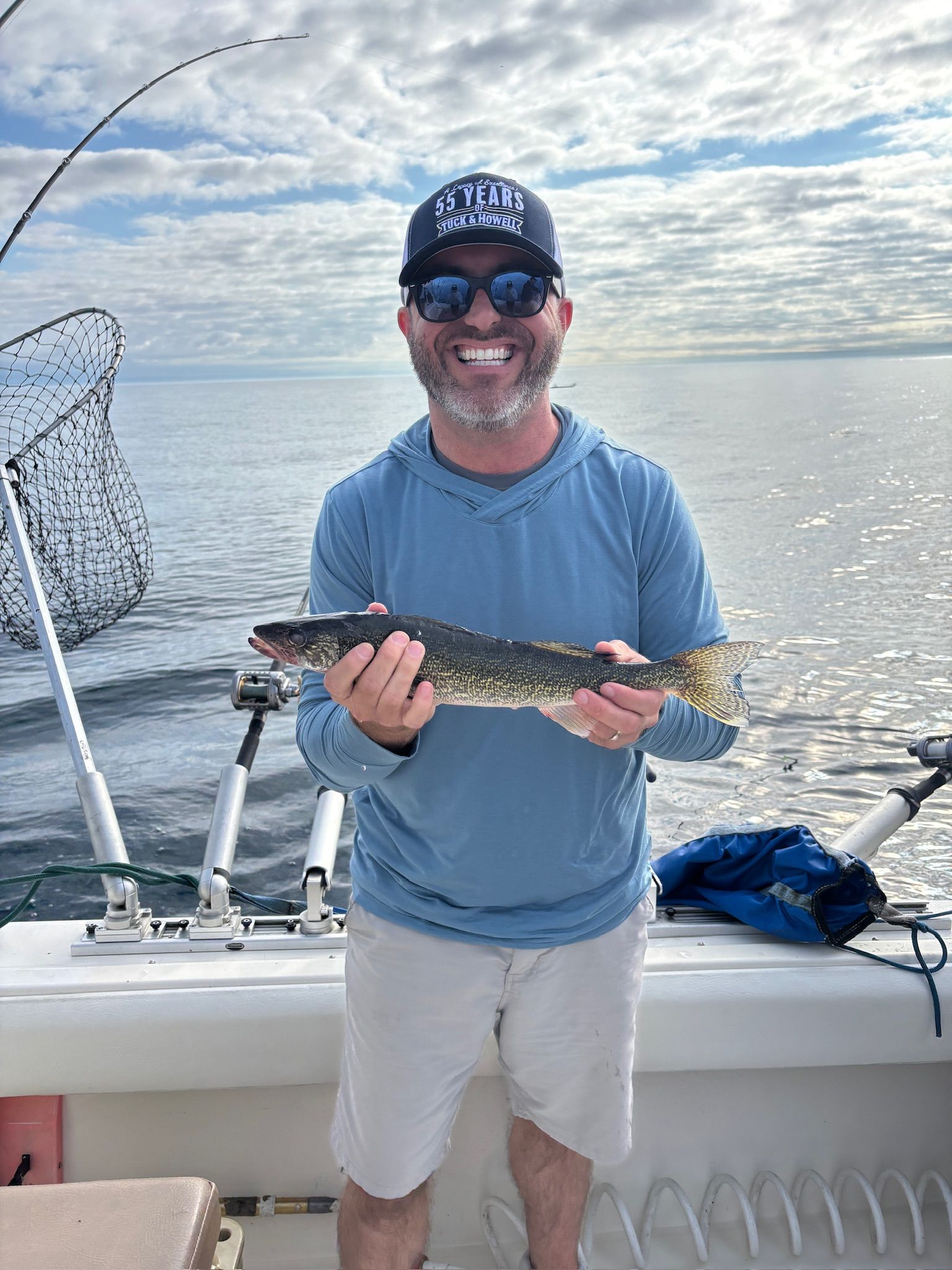Man smiles holding a fish on a boat, wearing sunglasses and a blue shirt, overlooking a lake.