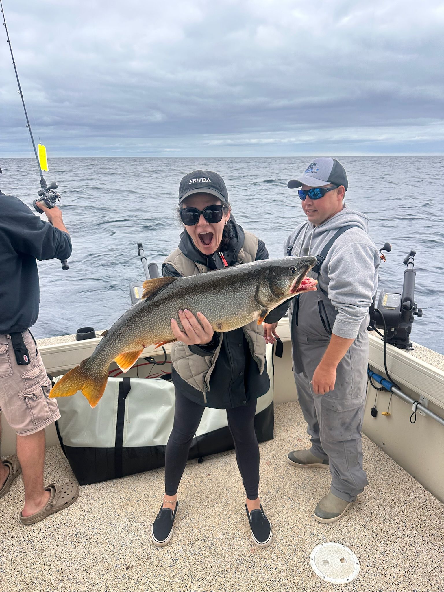 Woman excitedly holds a large fish on a boat, assisted by a man; cloudy day on the water.