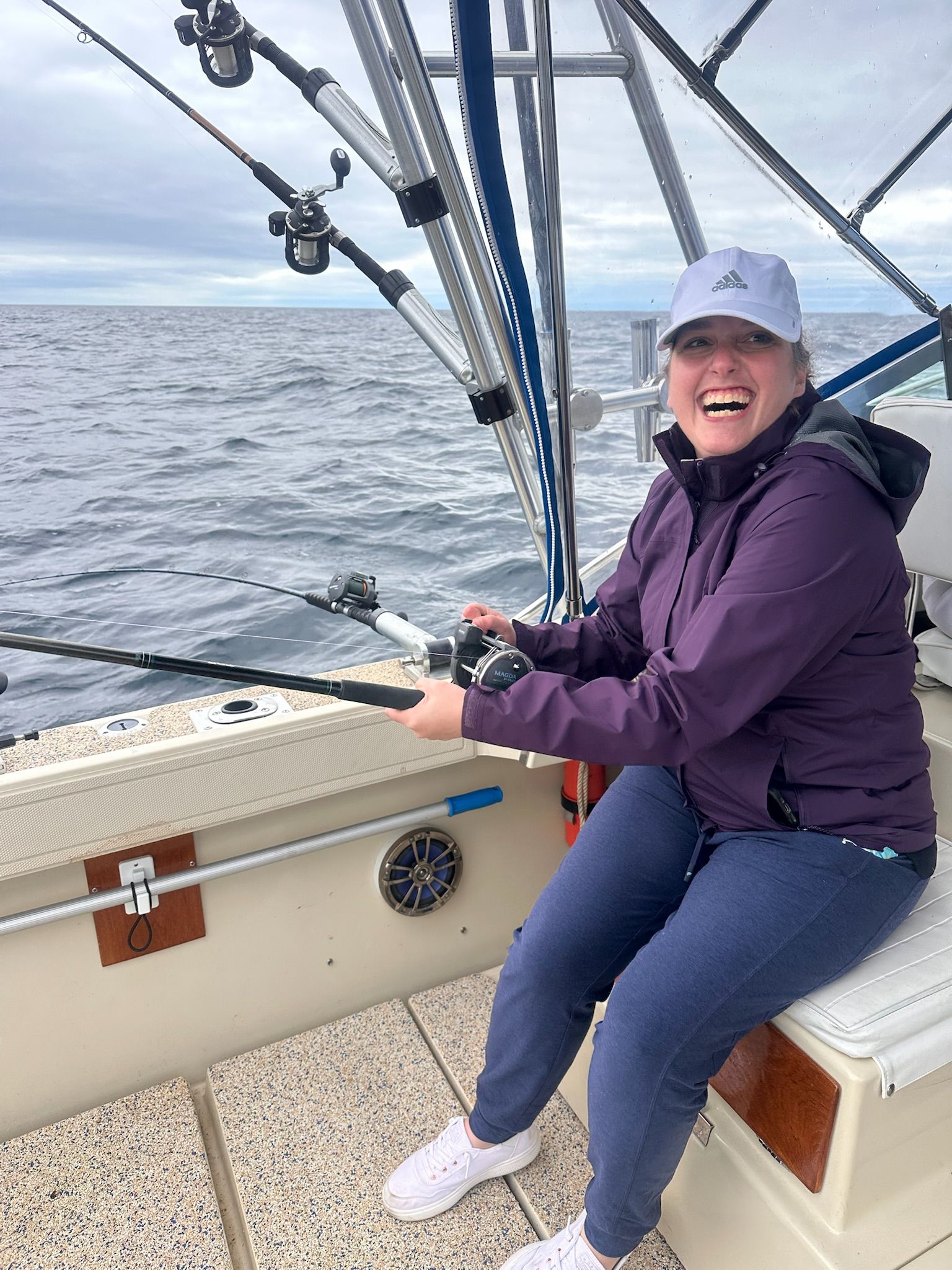 Woman fishing on a boat; laughing, wearing purple jacket and baseball cap, overcast sky.