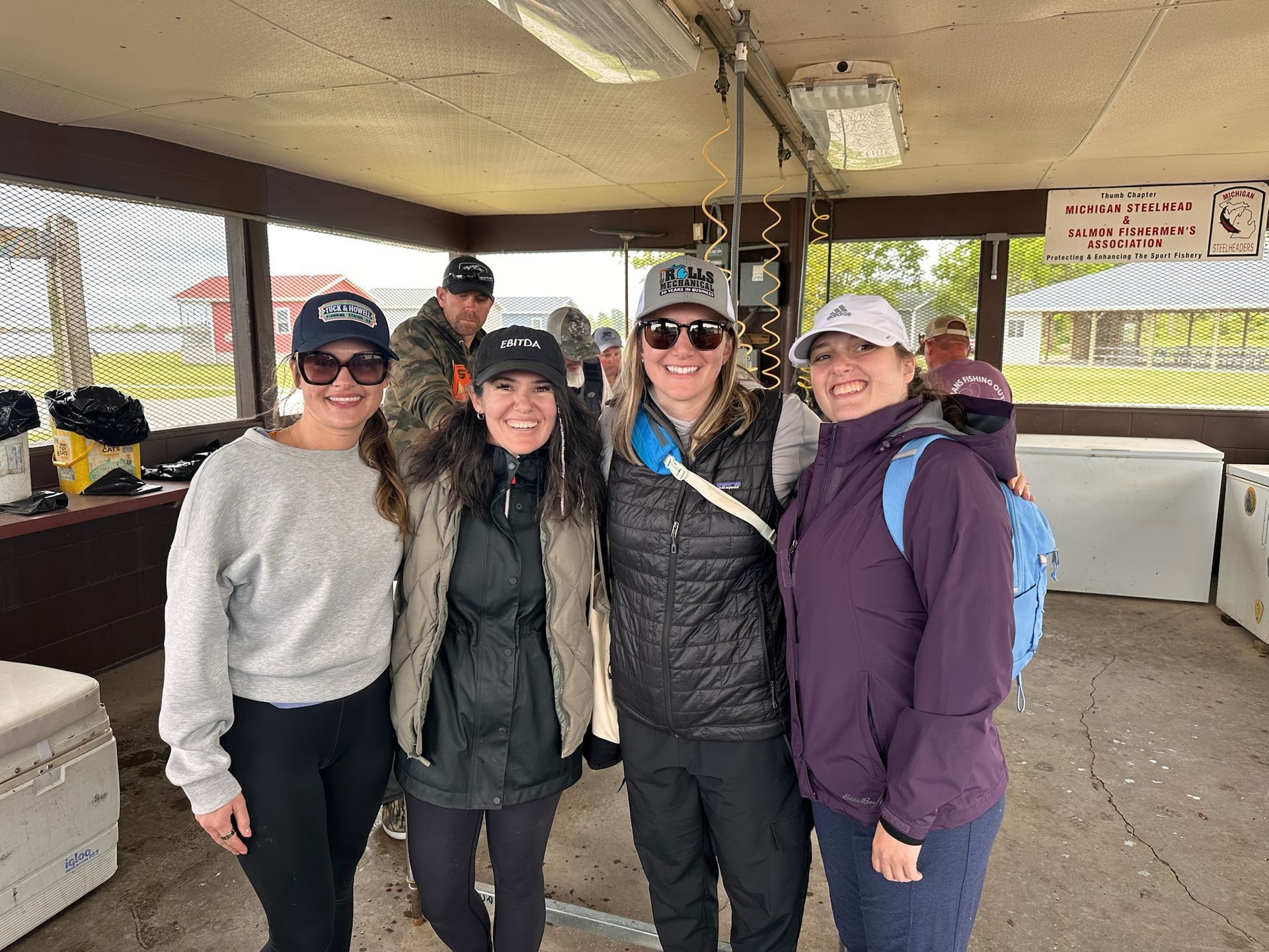 Four women smiling and posing under a shelter. They wear hats, sunglasses, and jackets. A man is in the background.