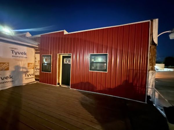 Red metal-sided building with black door and windows, Tyvek wrap, built on a deck at night.