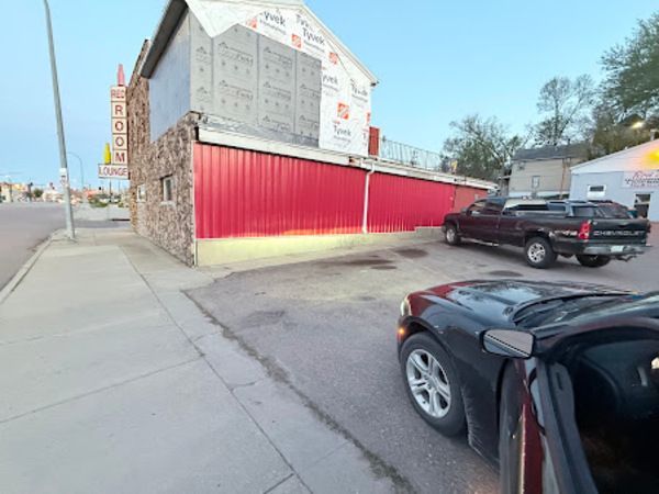 Exterior of a building with red siding, stone facade, and a sign. Two vehicles are parked nearby.
