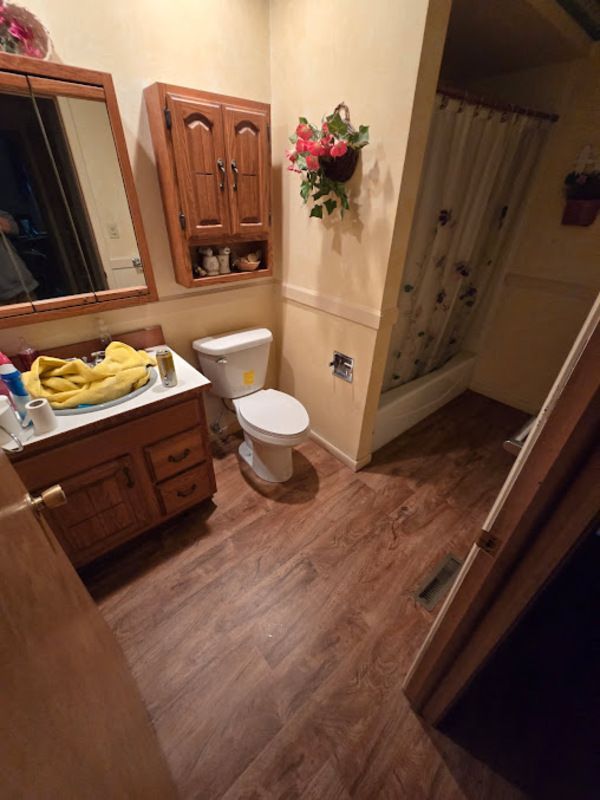 Bathroom with wood vanity, toilet, and tub/shower. Brown flooring and light yellow walls.