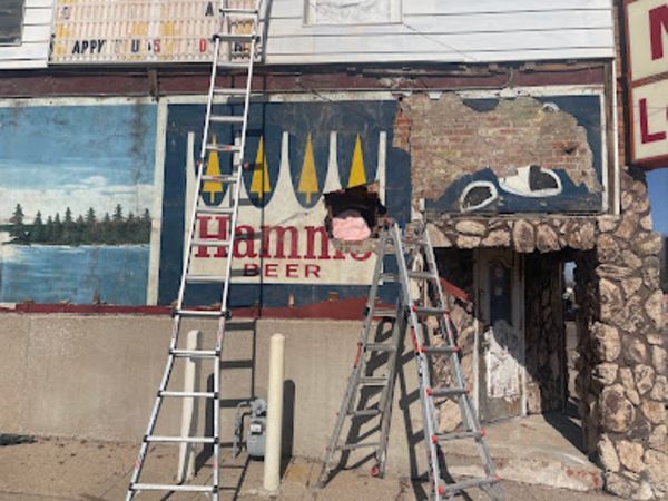 Exterior of a bar with ladders, old painted sign for Hamm's Beer, peeling paint, and stone facade.