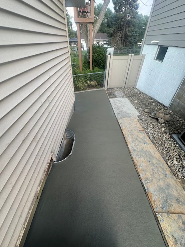 Freshly poured concrete sidewalk next to a house with tan siding and a white fence.