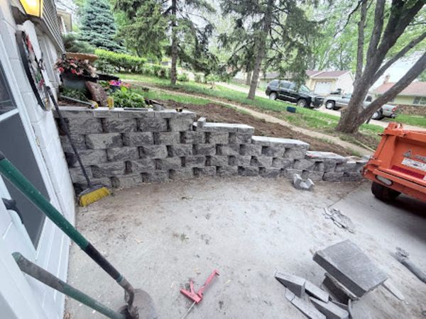 Retaining wall made of gray blocks on a sloping yard; tools, orange truck, and cars in the background.