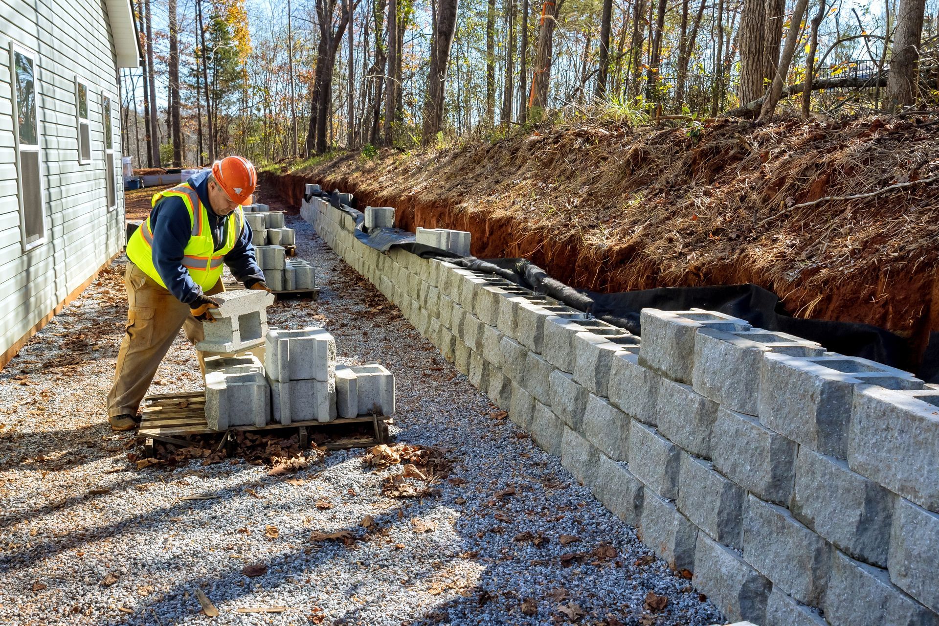 Construction worker building a retaining wall with concrete blocks outdoors near a building and trees.