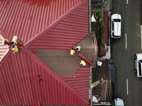 Roofers in yellow vests working on a red metal roof, aerial view. Cars parked on the street beside the house.