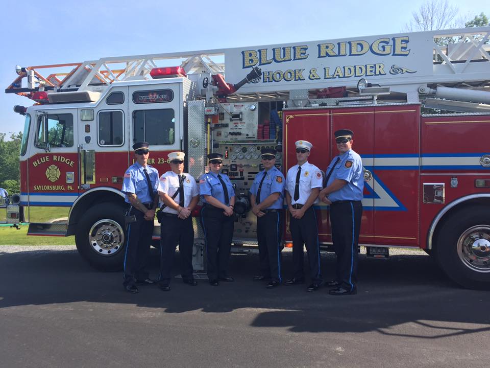 A group of firefighters stand in front of a blue ridge hook and ladders fire truck