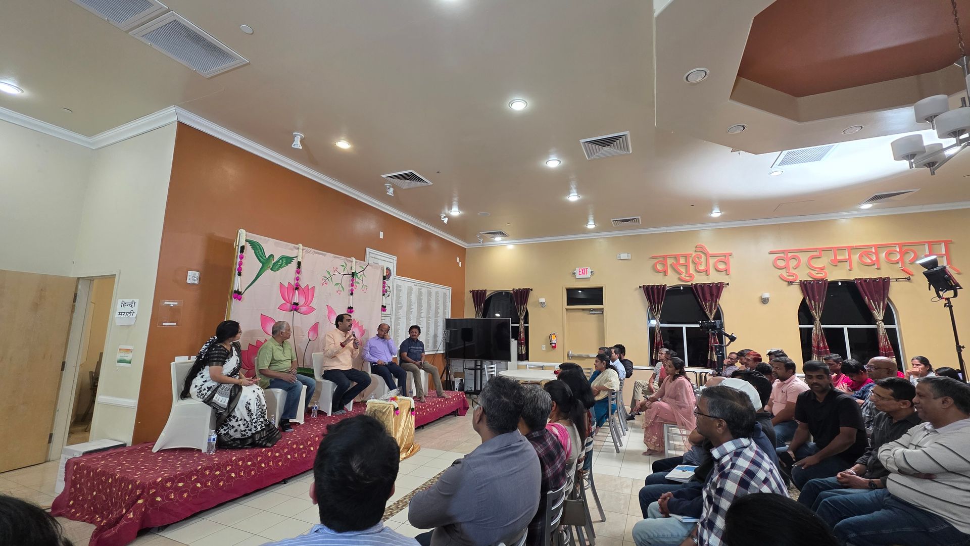 Audience watches a stage performance in a decorated hall with red drapes and floral backdrop.