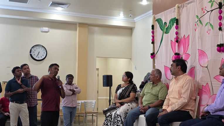 People seated and standing in a decorated room, with a man speaking to the group near a clock and wall speaker.