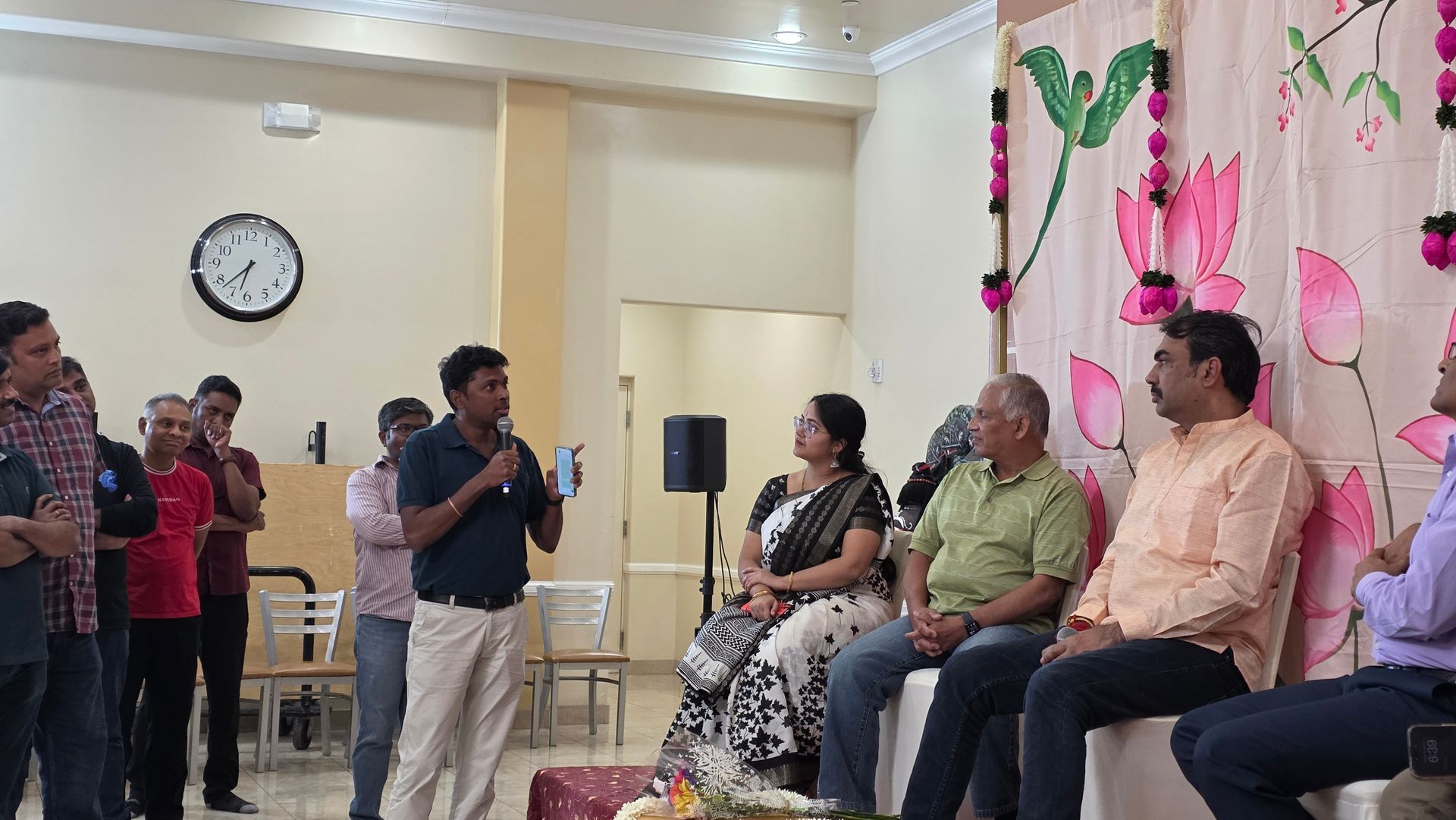 People seated in a decorated room while a man speaks into a microphone.