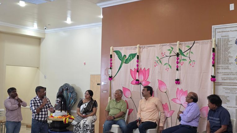 People seated on a stage in a hall, with a pink floral backdrop and a microphone at the left.