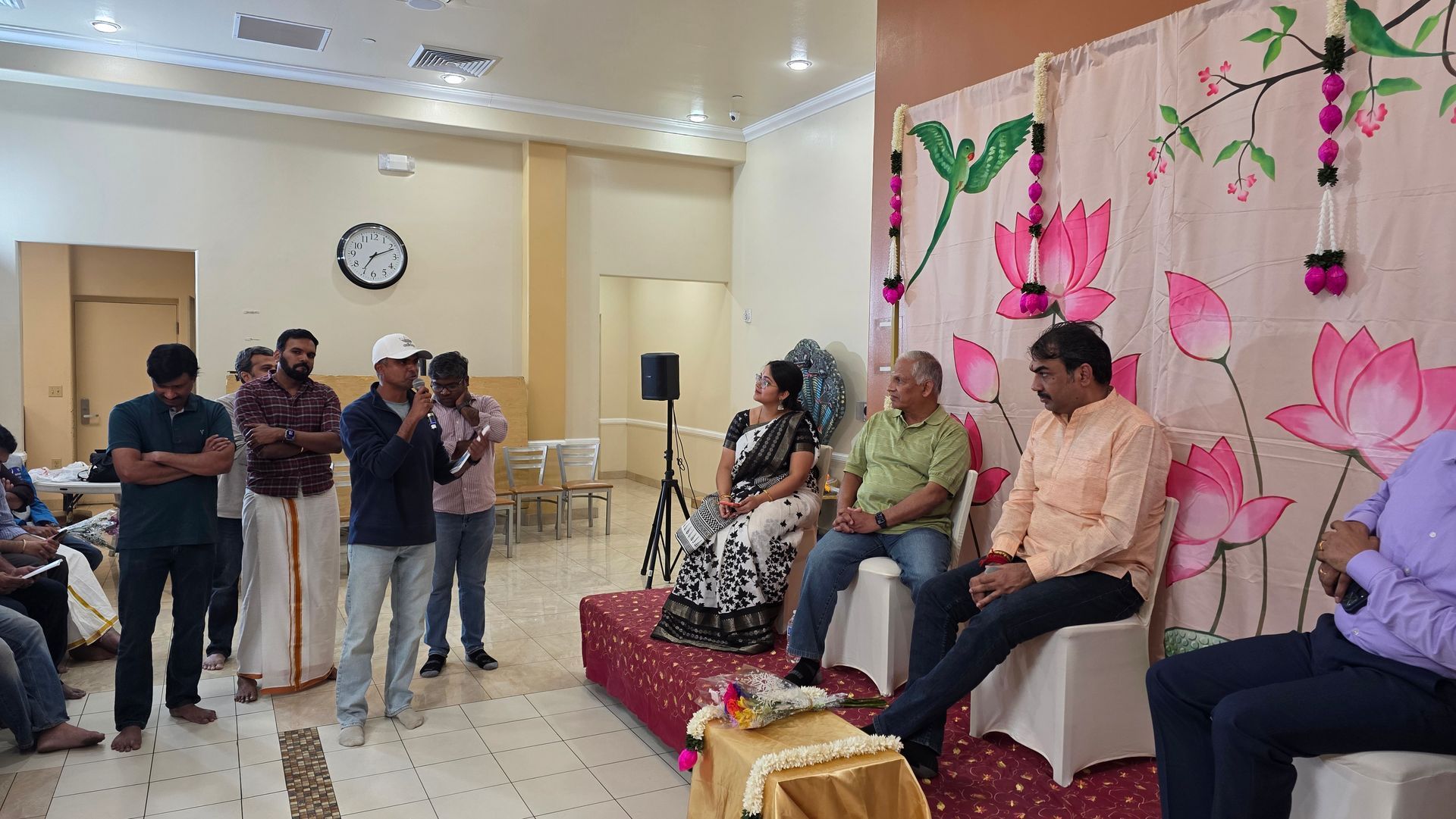 People seated on a stage with floral backdrop while photographers stand nearby in a hall