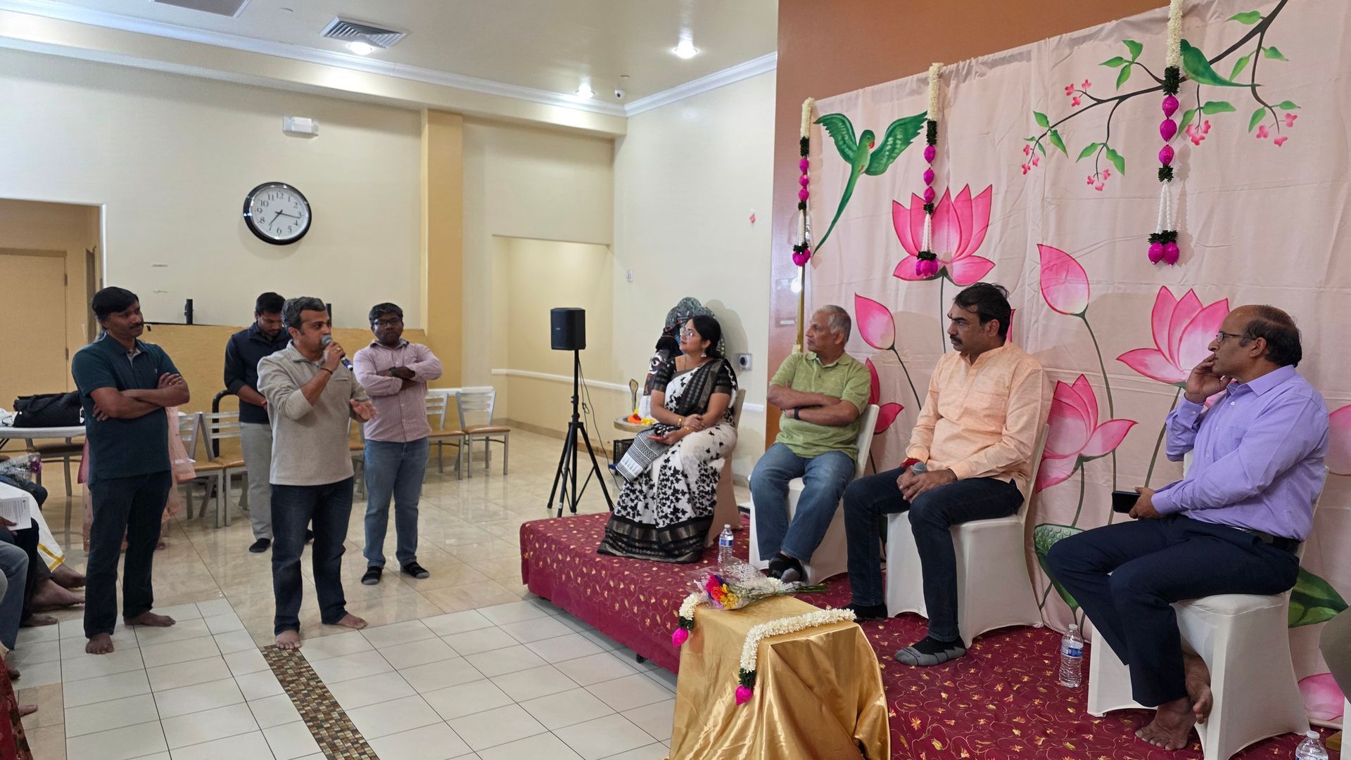 People seated on a decorated stage during an indoor gathering, with others standing nearby and a wall clock in back