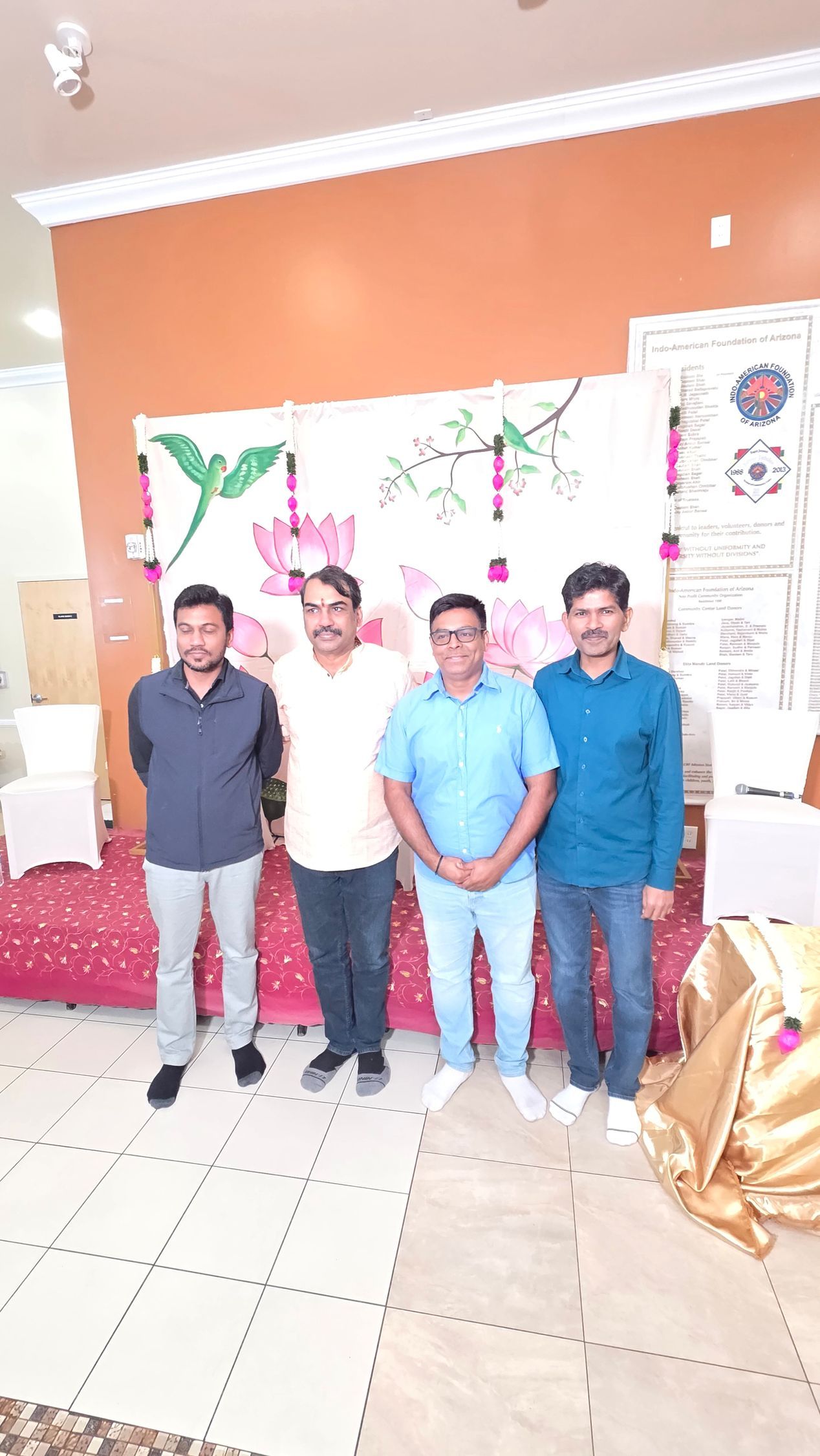 Four people standing indoors in front of a decorated orange wall and banner, posing for a photo.