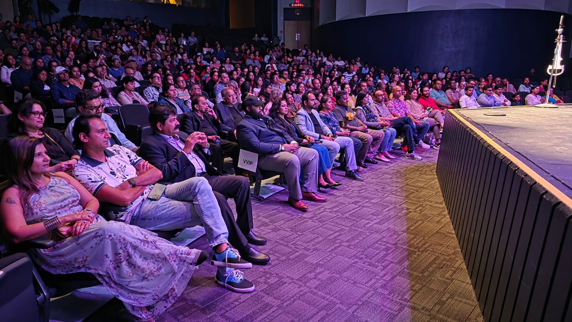 Audience seated in a dim auditorium, watching a stage lit in purple and pink