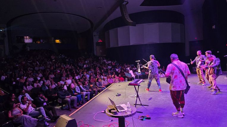Band performing on a purple-lit stage before a large seated audience in a concert hall.