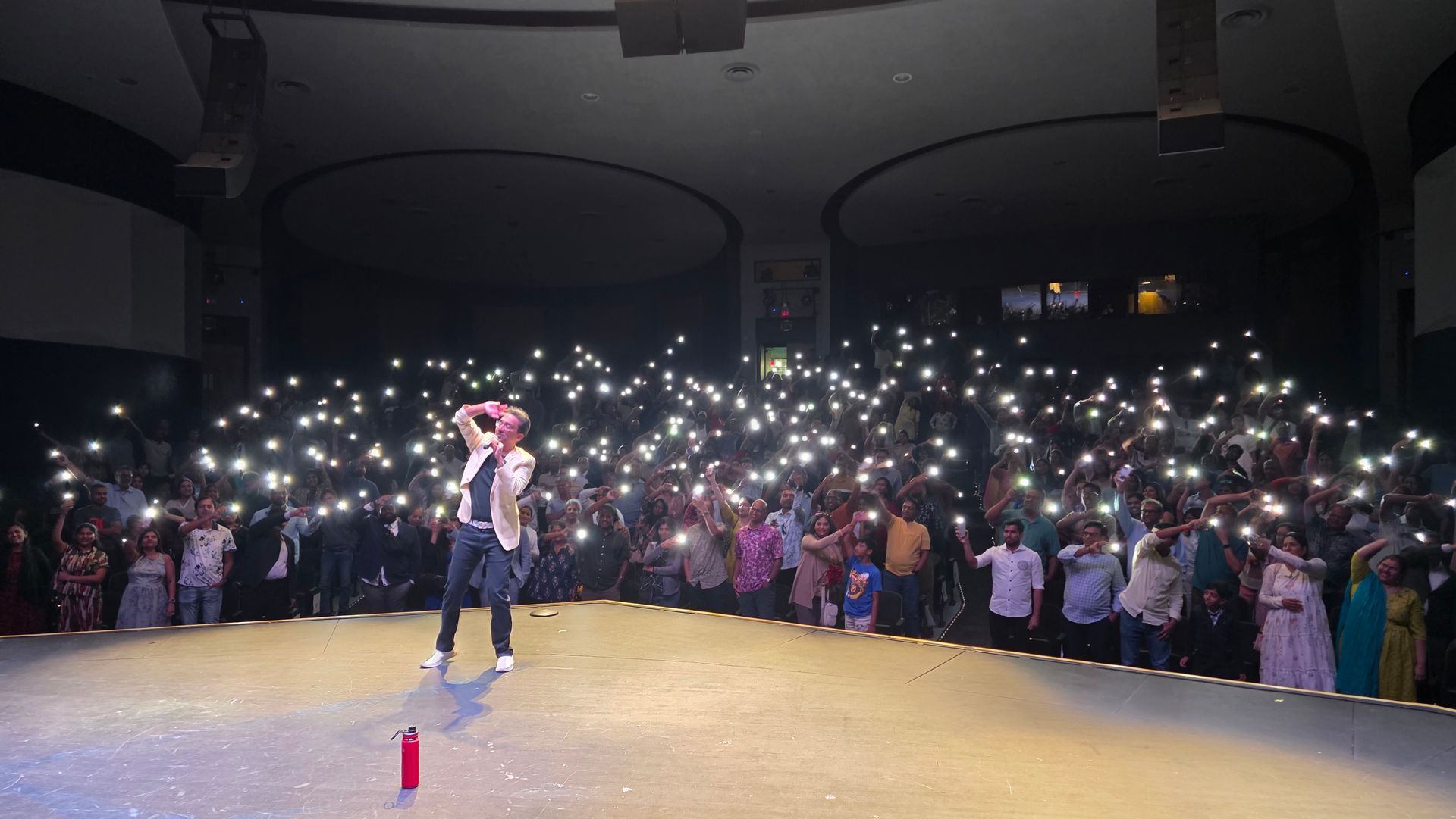 Performer on stage facing a crowd holding up phone flashlights in a dark auditorium