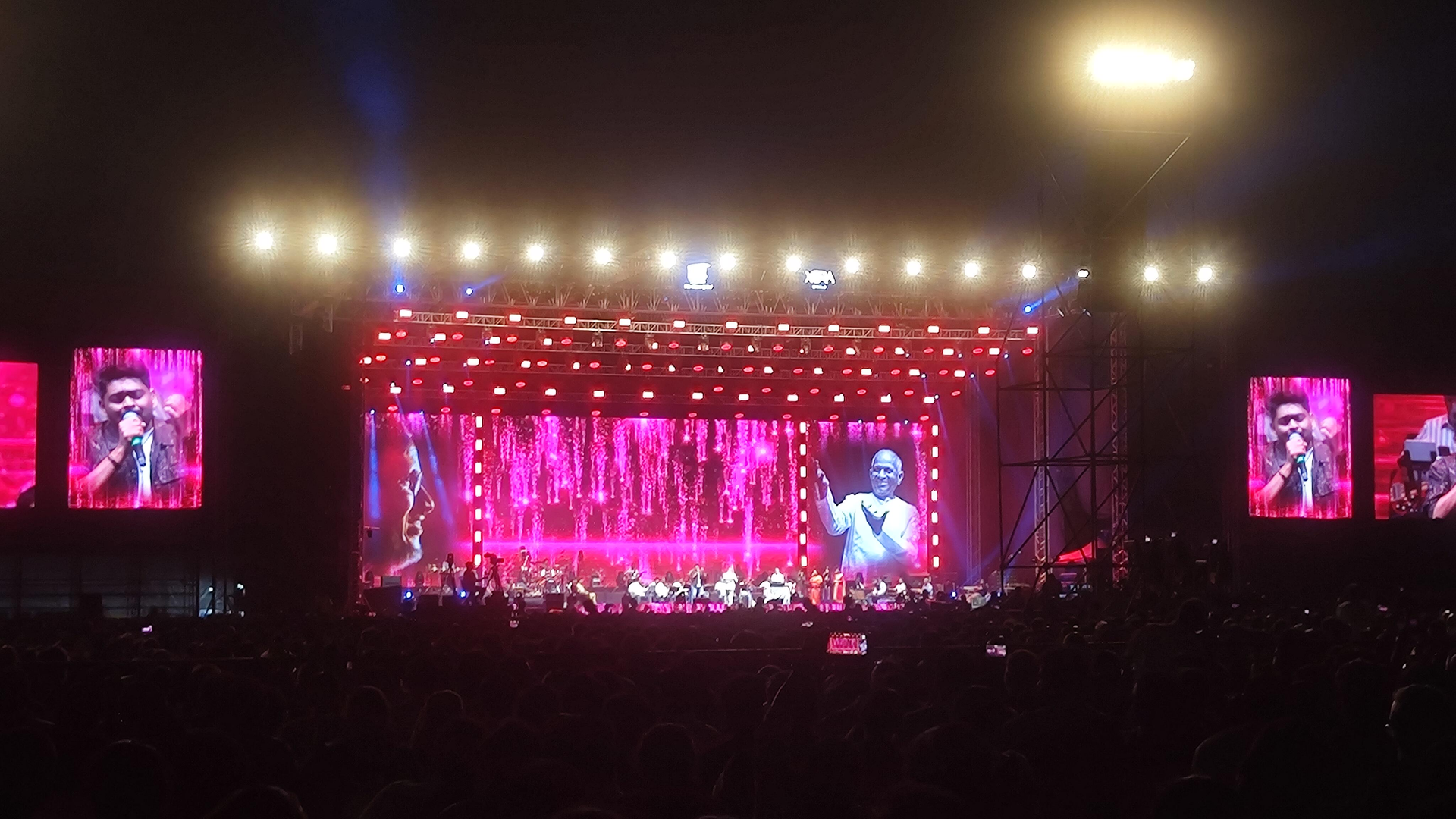 Nighttime concert stage with bright pink lights and giant screens, viewed from the crowd