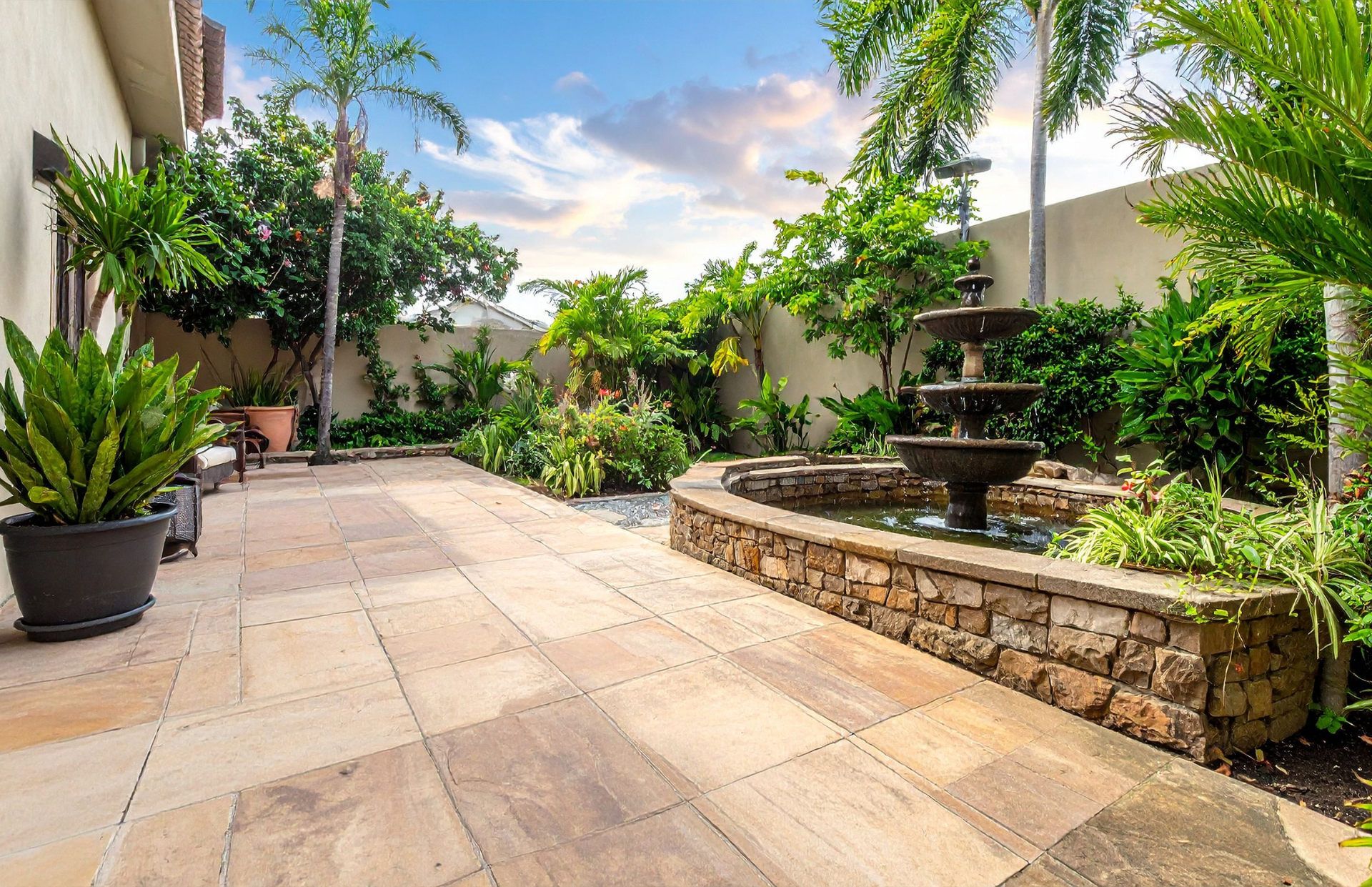 Sunlit patio with stone pavers, lush plants, and a small fountain beside a stucco wall
