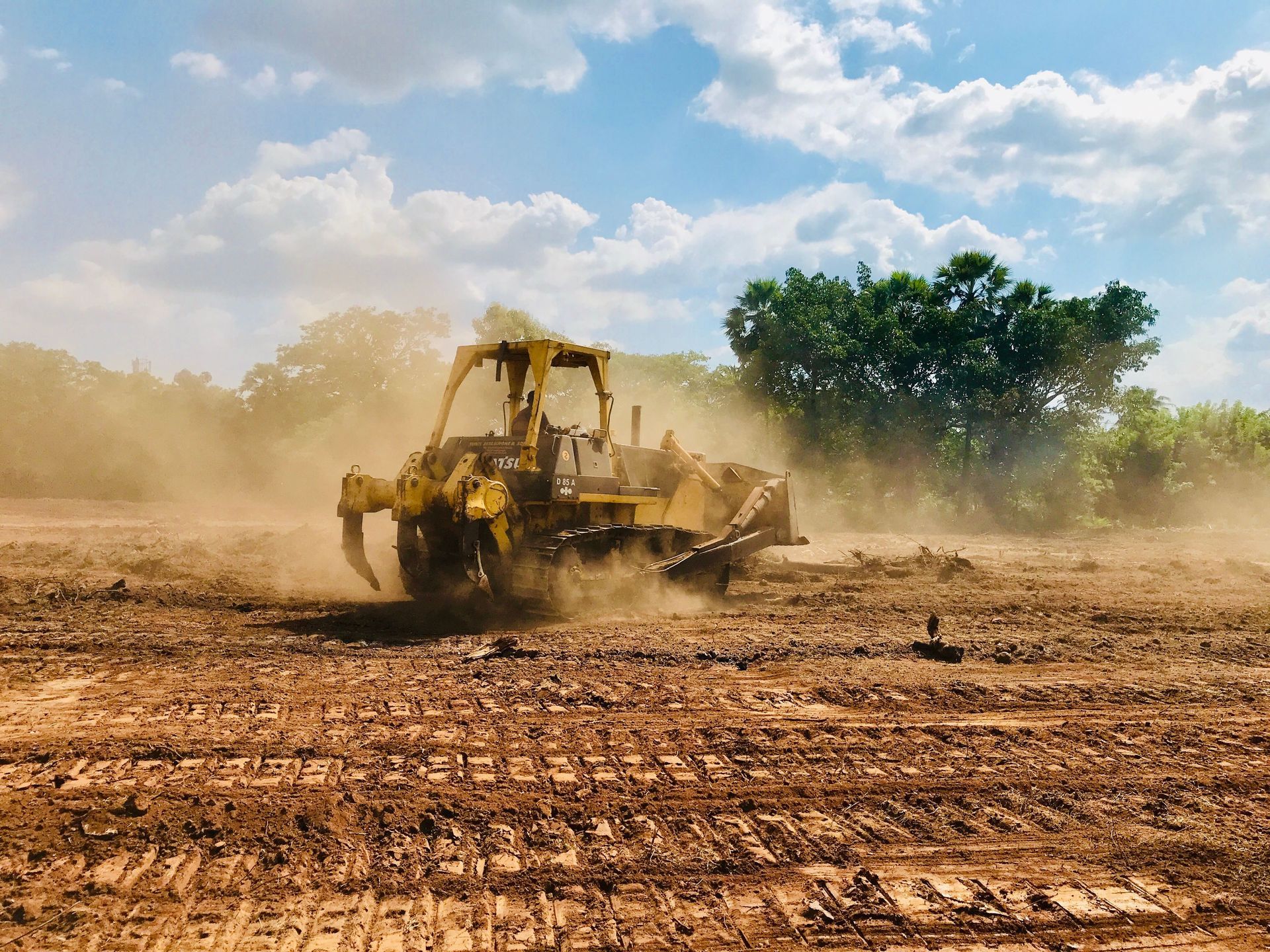 Yellow bulldozer grading a dusty field under a blue sky with trees in the background