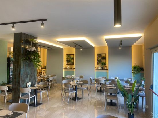 Interno di un ristorante moderno con tavoli, sedie e piante su pareti grigie e gialle.