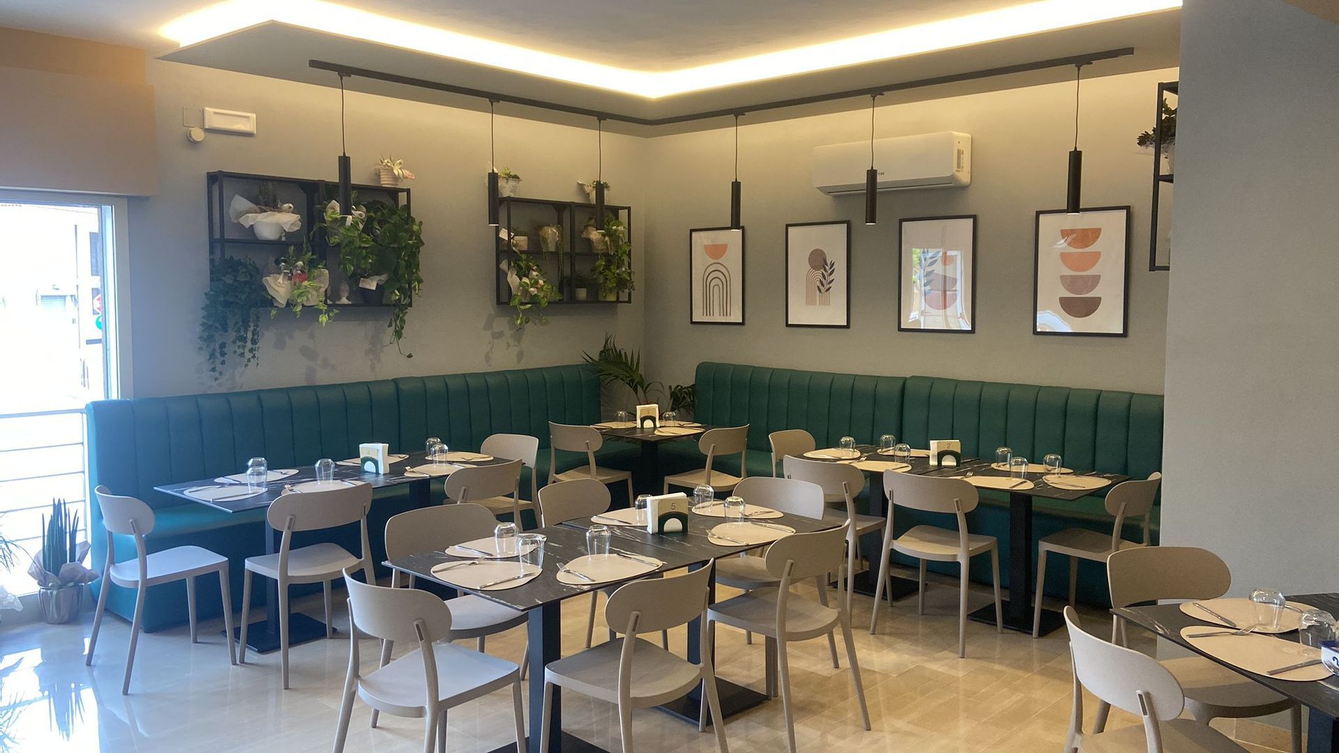Interno del ristorante: panche verdi, tavoli con apparecchiati, sedie grigio chiaro, opere d'arte, piante e porta aperta.