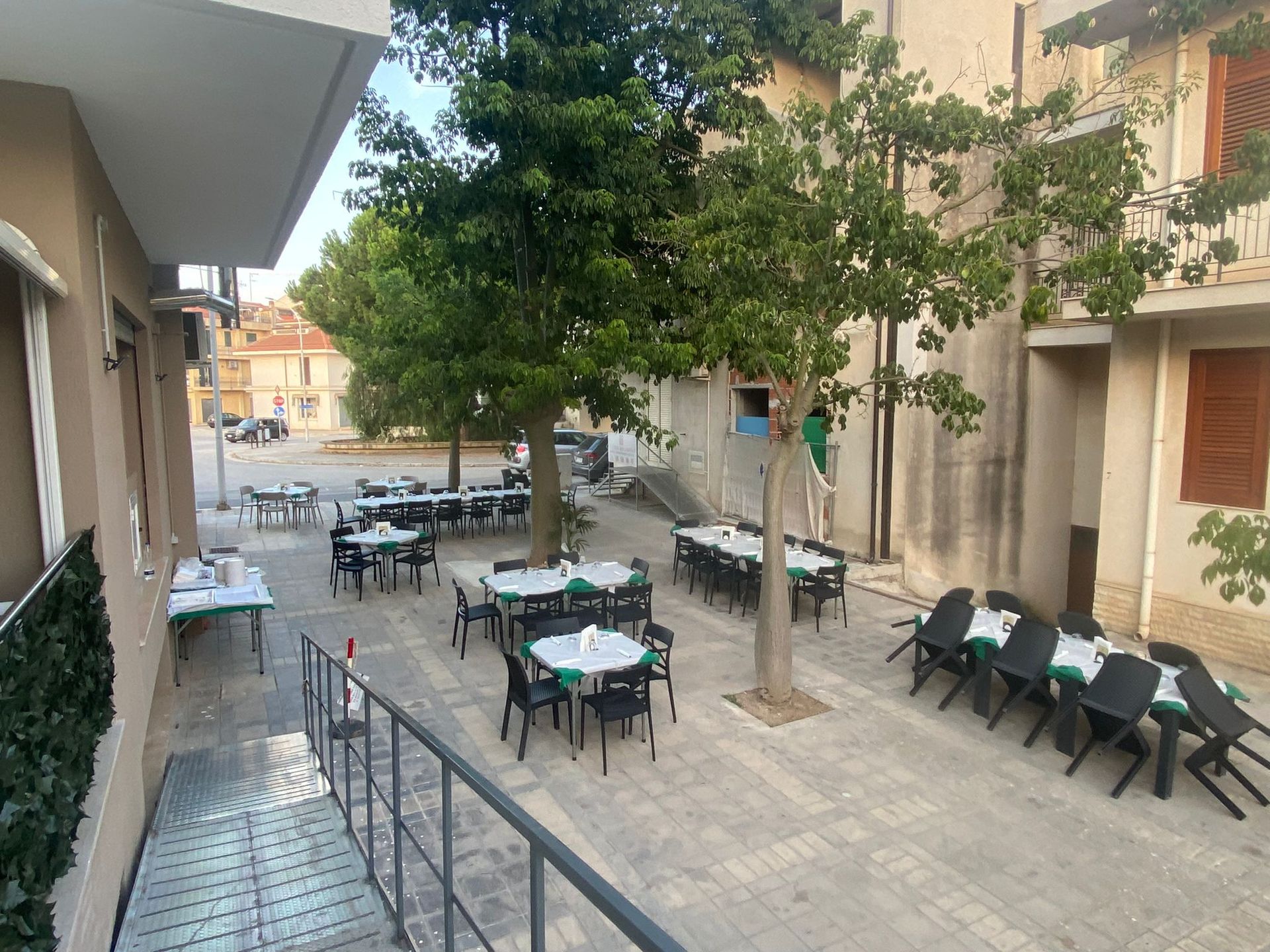 Tavoli e sedie all'aperto sistemati in una piazza pavimentata, probabilmente per un ritrovo. Tovaglie verdi. Edifici circostanti.