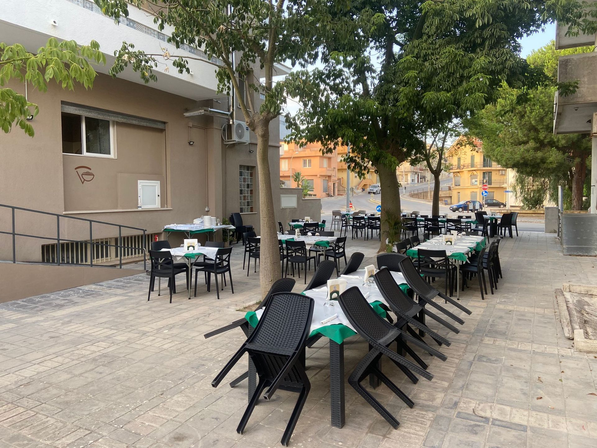 Ristorante all'aperto con sedie nere disposte su tovaglie verdi sotto gli alberi.