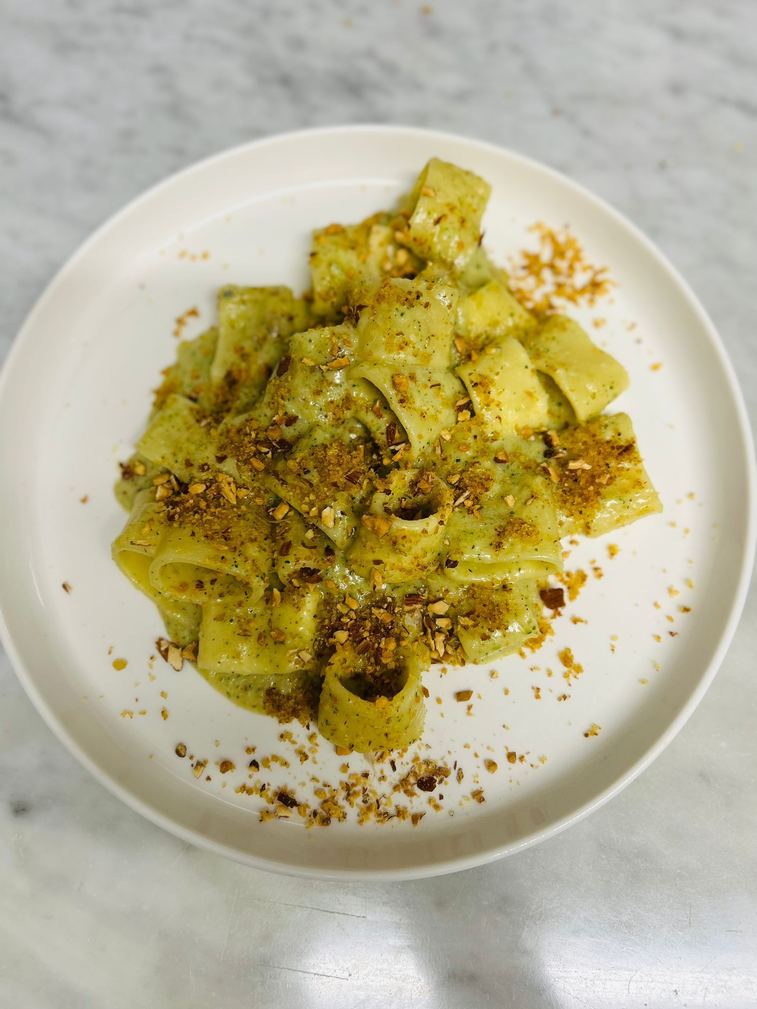 Pasta con salsa verde e condimento croccante su un piatto bianco.