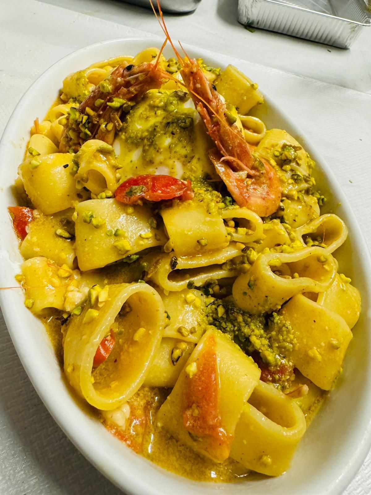 Piatto di pasta con pesto verde, pomodori, gamberi e pistacchi, in una ciotola bianca ovale.