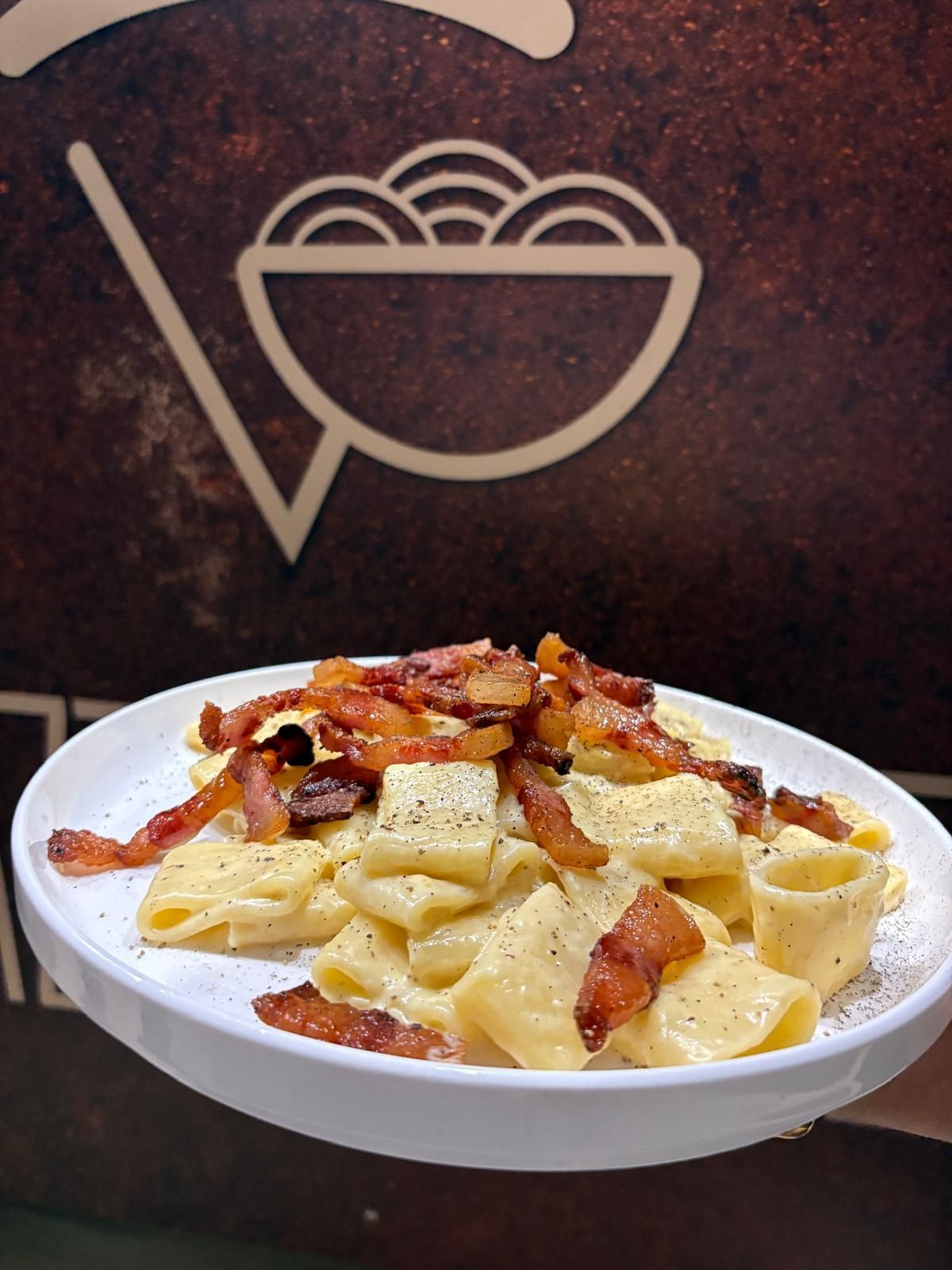 Piatto di pasta cremosa con pancetta croccante, davanti a un logo alimentare.