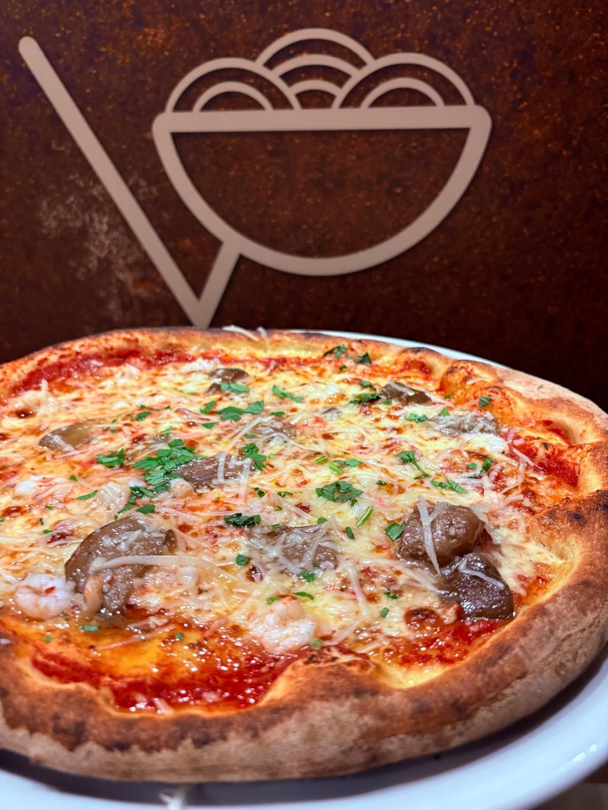 Pizza con formaggio, funghi e prezzemolo su un piatto bianco, con il logo del ristorante sullo sfondo.