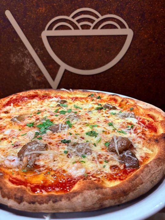 Pizza con formaggio, funghi e prezzemolo su un piatto bianco, con il logo del ristorante sullo sfondo.