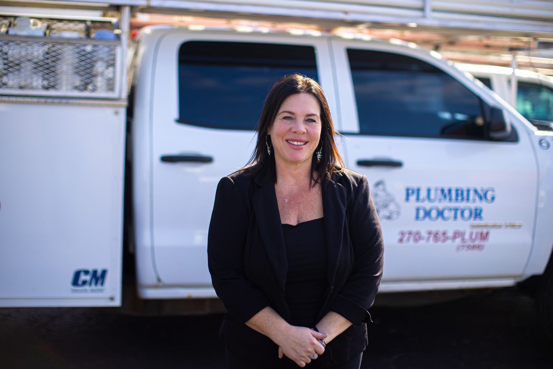 Woman in black blazer smiles in front of a white Plumbing Doctor truck.