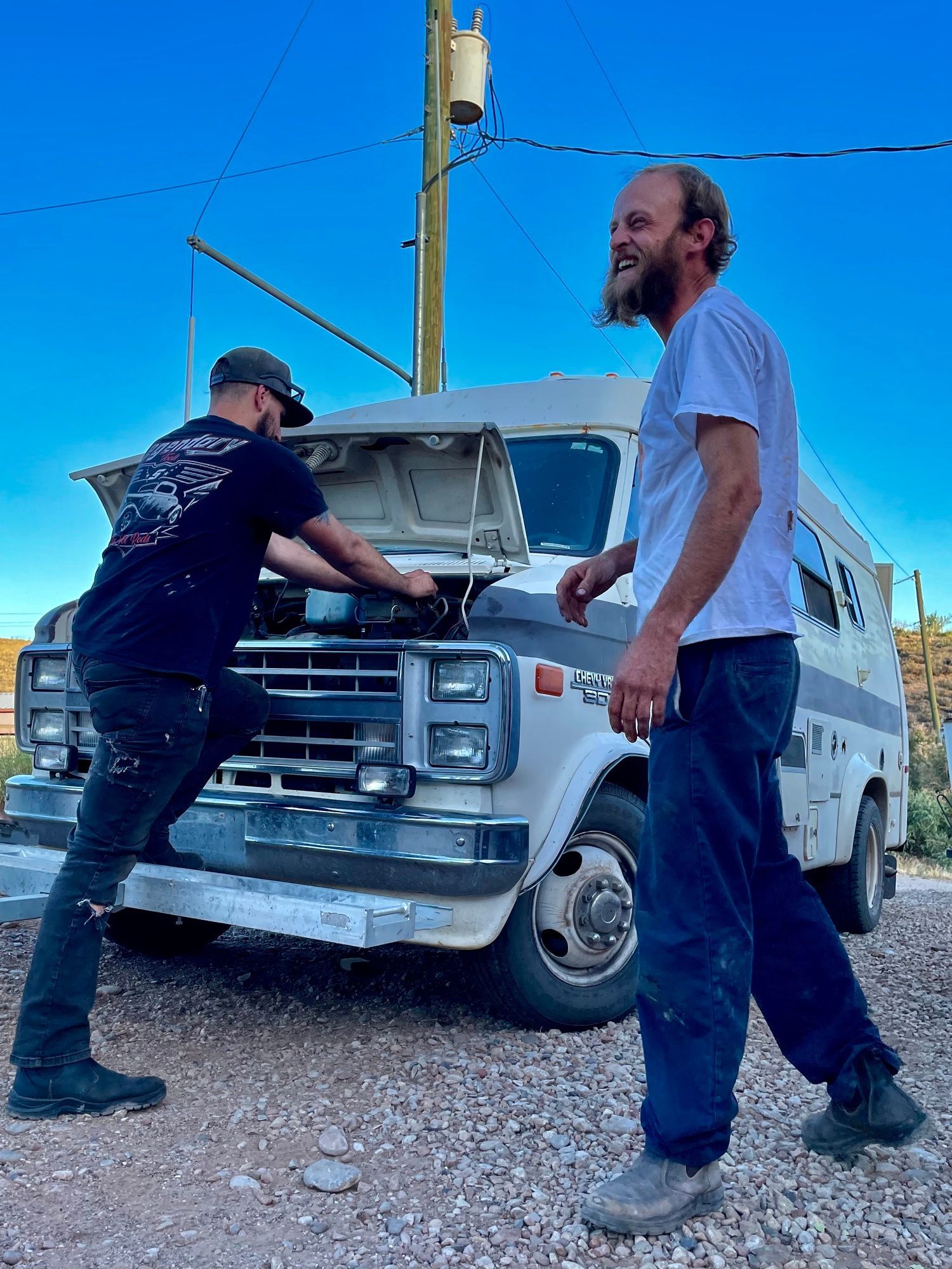 Two men are working on a van in a gravel lot.