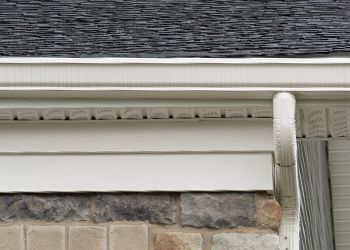 White gutter and downspout on a house with a gray roof and stone wall.