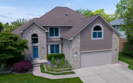 Two-story suburban home with stone and brown siding, a double garage, and a manicured front lawn under a blue sky.