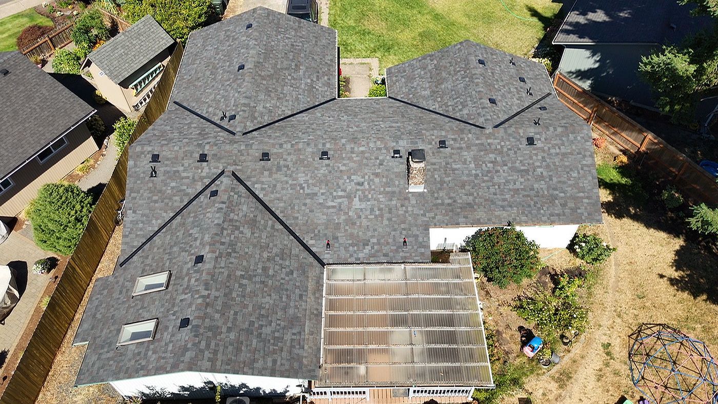 An aerial view of a dark-shingled residential roof with a rectangular patio cover and surrounding landscaping.