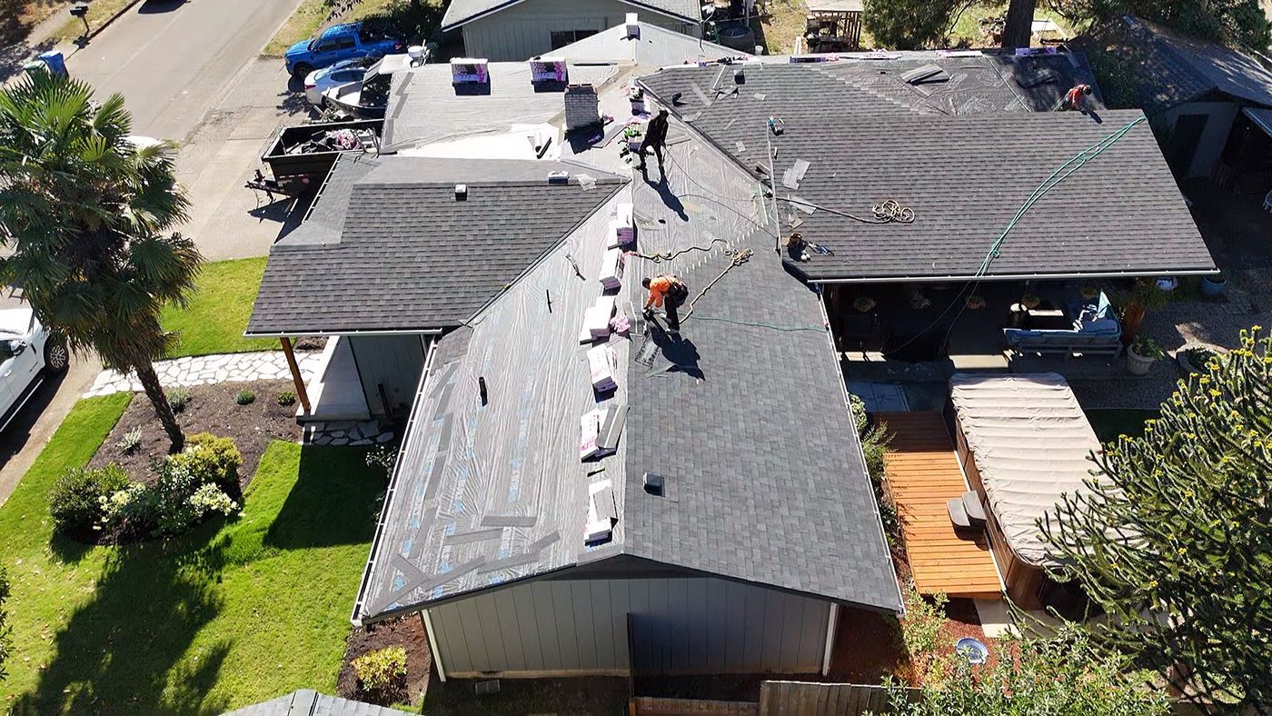 An aerial view of two workers installing gray asphalt shingles on the roof of a suburban house.