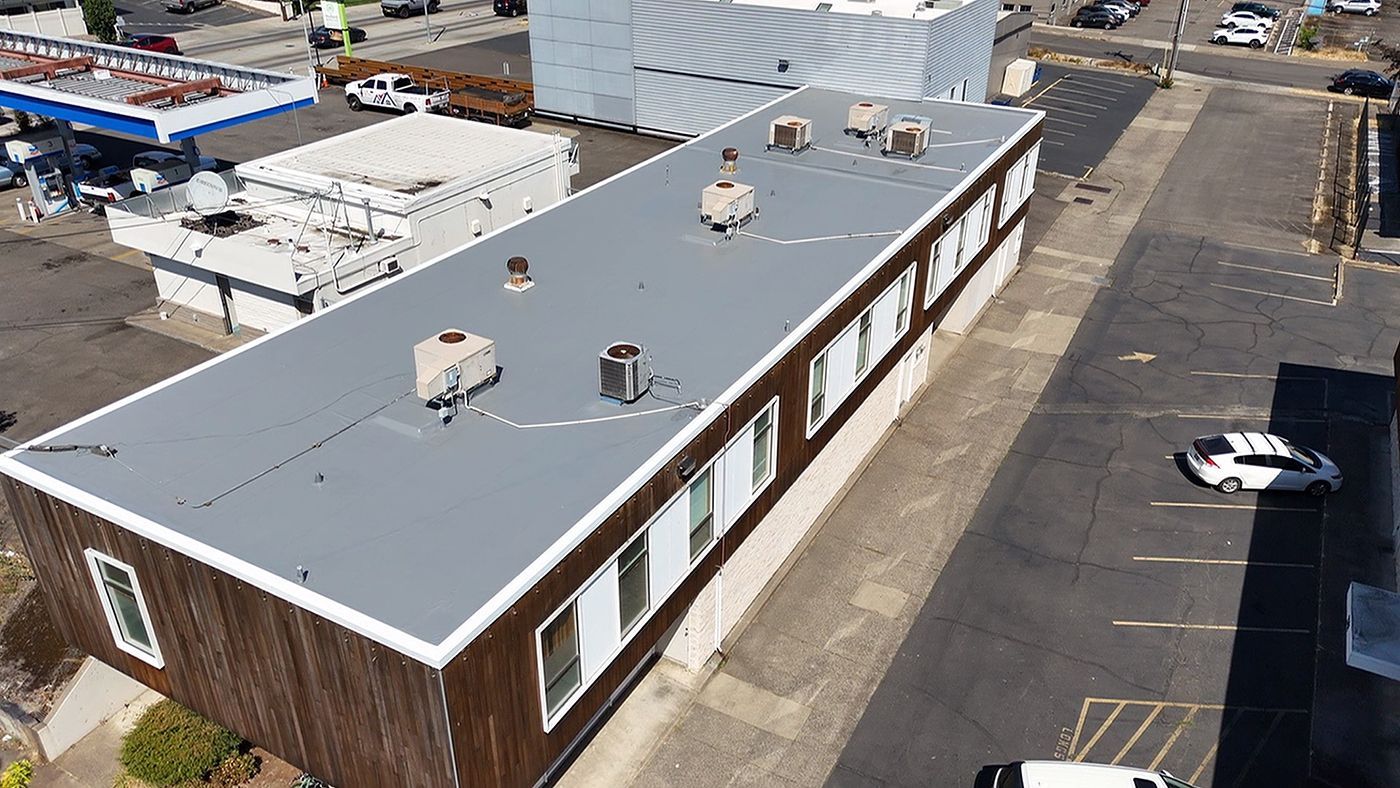Aerial view of a long, low, brown building with a flat gray roof and windows, parked cars on the street.