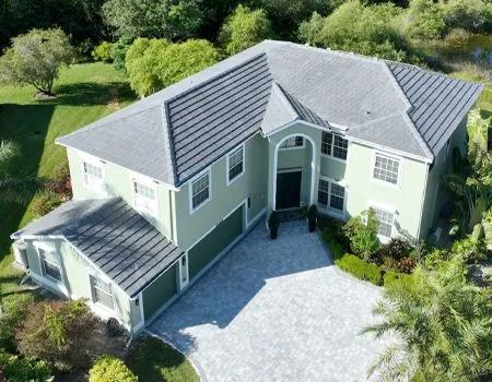Two-story house with green exterior, gray roof, and stone driveway surrounded by green grass and trees.