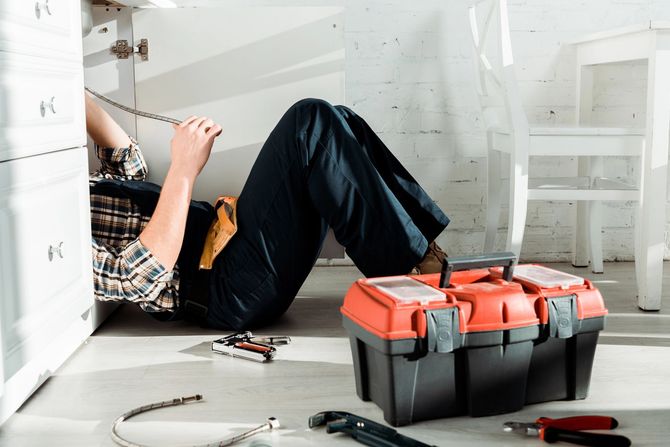 A person in work clothes kneeling under a kitchen sink to perform repairs, with a toolbox and tools nearby on the floor.
