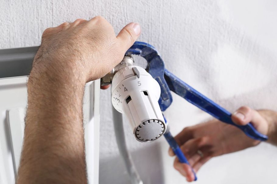 Hands use blue pliers to tighten or remove a white thermostatic radiator valve on a radiator against a white wall.