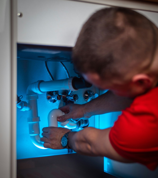 A plumber in a red shirt works on white PVC pipes under a sink illuminated by blue light.
